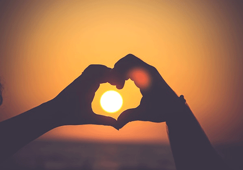 pessoas fazendo um coração com as mãos no pôr do sol com o mar no fundo